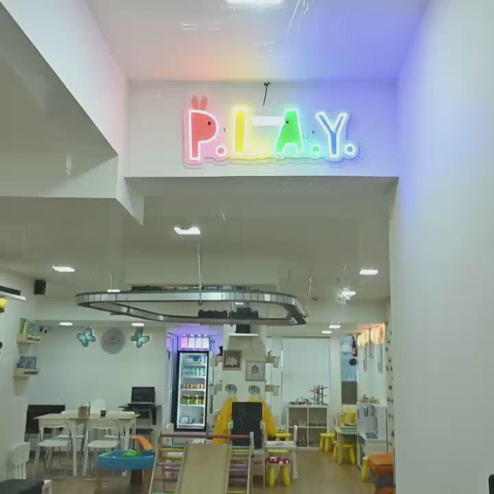 This vibrant play space, likely for young children, features a colorful "P.L.A.Y." sign, a miniature train track, a slide, and a world map mural. The environment is bright and engaging, with shelves stocked with books and toys, creating a fun and stimulating atmosphere.