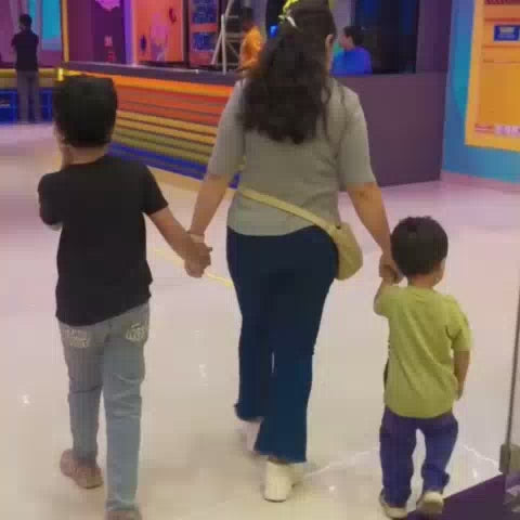 Children and adults enjoy a vibrant indoor trampoline park filled with colorful foam pits, slides, and climbing walls. The atmosphere is energetic and playful, with people of various ages bouncing, climbing, and sliding. The park features bright neon lights and a lively, fun-filled environment.