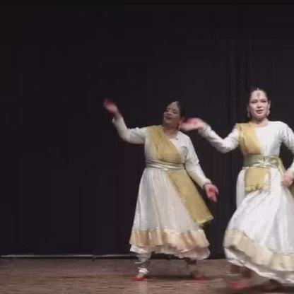 Three women in traditional white and gold Indian dance attire perform a graceful Kathak dance on a wooden stage with a black backdrop. Their synchronized movements and flowing skirts create a visually captivating performance.