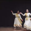 Three women in traditional white and gold Indian dance attire perform a graceful Kathak dance on a wooden stage with a black backdrop. Their synchronized movements and flowing skirts create a visually captivating performance.