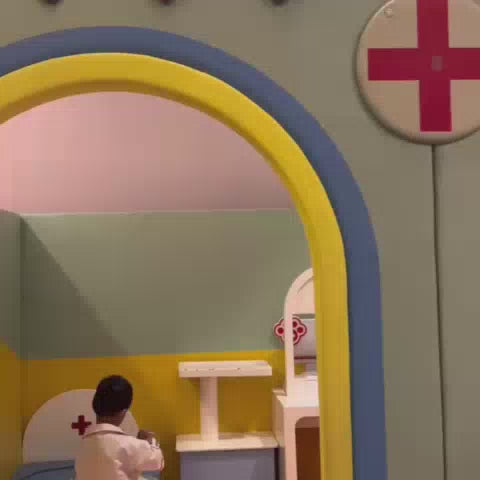 A young child enjoys a vibrant indoor play area with various themed zones, including a doctor's clinic, supermarket, and ball pit. The atmosphere is playful and engaging, with colorful equipment and interactive activities designed for children's exploration and fun. Parents can relax while their children engage in imaginative play and physical activities.