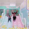 A young child and an adult enjoy a vibrant indoor play area filled with colorful slides, ball pits, and interactive stations. The playful environment features a "Welcome" sign, a "Fun Block" cafe, and various engaging activities like a pin screen and a water play area. The atmosphere is cheerful and energetic, perfect for children to explore and have fun.