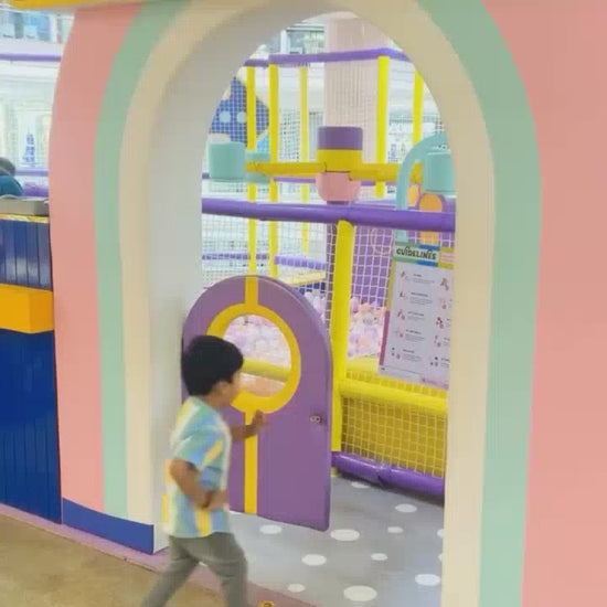 A young child joyfully navigates a colorful indoor playground filled with soft balls and playful equipment. The environment is bright and engaging, designed for toddlers and young children to explore and have fun. The child interacts with various attractions, including a ball pit, a mini kitchen, and swings, creating a lively and happy atmosphere.