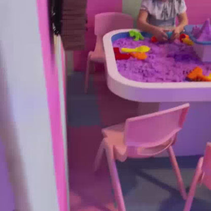 Children of various ages enjoy a vibrant indoor play area filled with colorful ball pits, slides, and a kinetic sand table. The atmosphere is lively and playful, with kids engaging in activities like sliding, jumping on a trampoline, and building with sand. Soft, pastel-colored structures and walls create a cheerful and inviting environment.