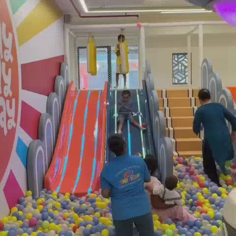 Children of various ages enjoy a vibrant indoor play area filled with colorful ball pits, slides, and climbing structures. The atmosphere is lively and fun, with parents supervising and participating in the activities. The space features engaging attractions like a ball pit, a climbing wall, and a tube maze.