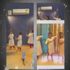In a dance studio, young children and adults practice Indian classical dance. The atmosphere is focused and energetic, with participants wearing traditional white attire. Equipment like an air conditioner is visible on the wall.