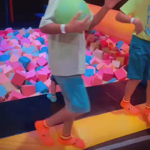 Children and adults enjoy a vibrant indoor trampoline park with a foam pit, climbing walls, and a zip line. The atmosphere is energetic and playful, with colorful equipment and neon lighting creating a fun environment for all ages.