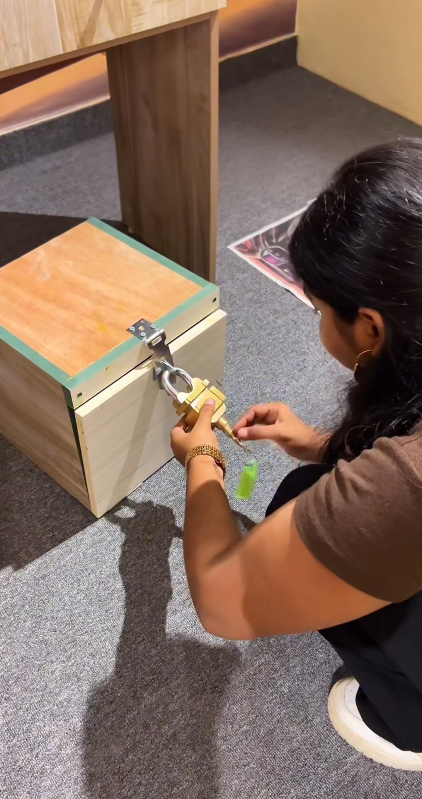 Person unlocking a wooden box with a large padlock while crouching on carpeted floor at Rajinder Nagar escape room