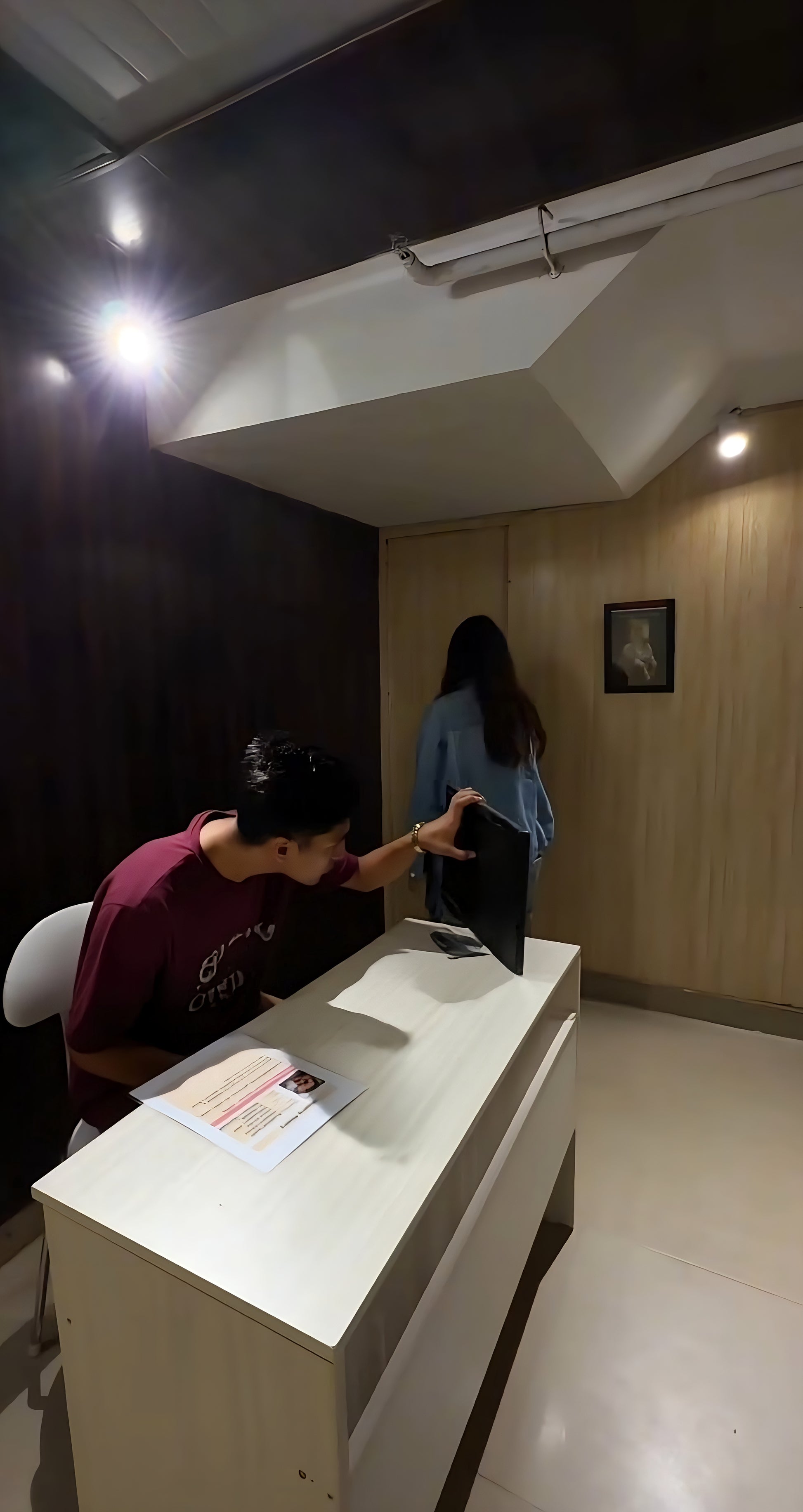 Person sitting at desk looking at monitor while woman walks toward wooden wall with framed picture in dim room