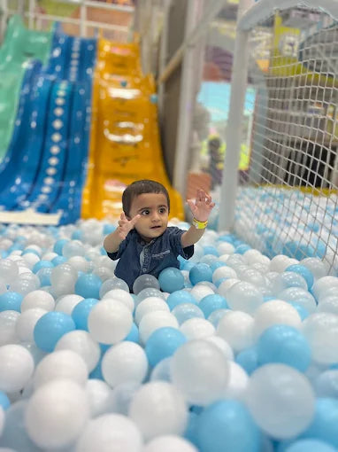 Young child playing in blue and white ball pit with colorful slides in the background at indoor trampoline park